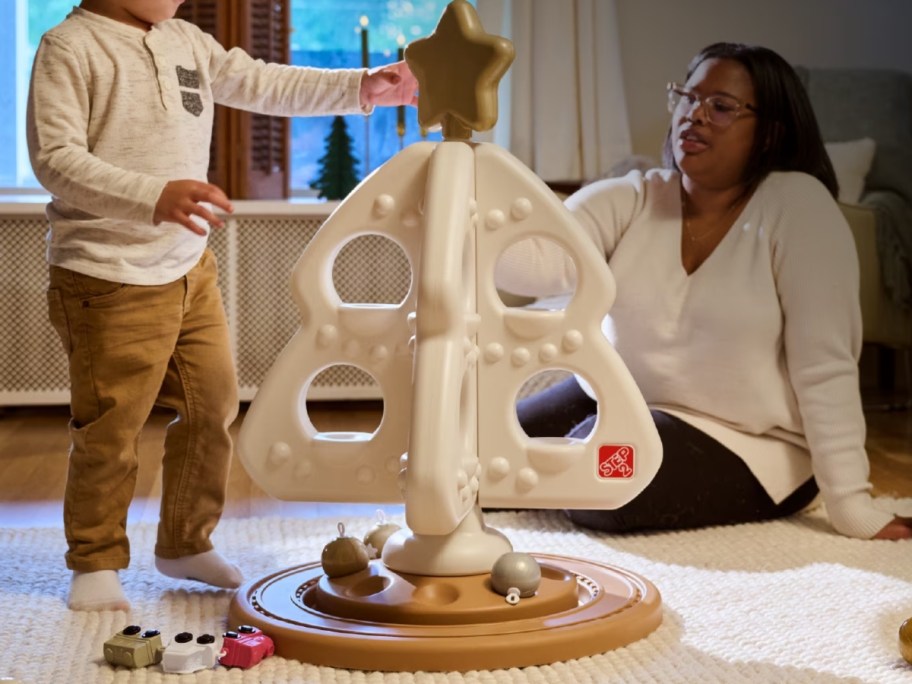 child adding star to top of white toy christmas tree while mom looks on